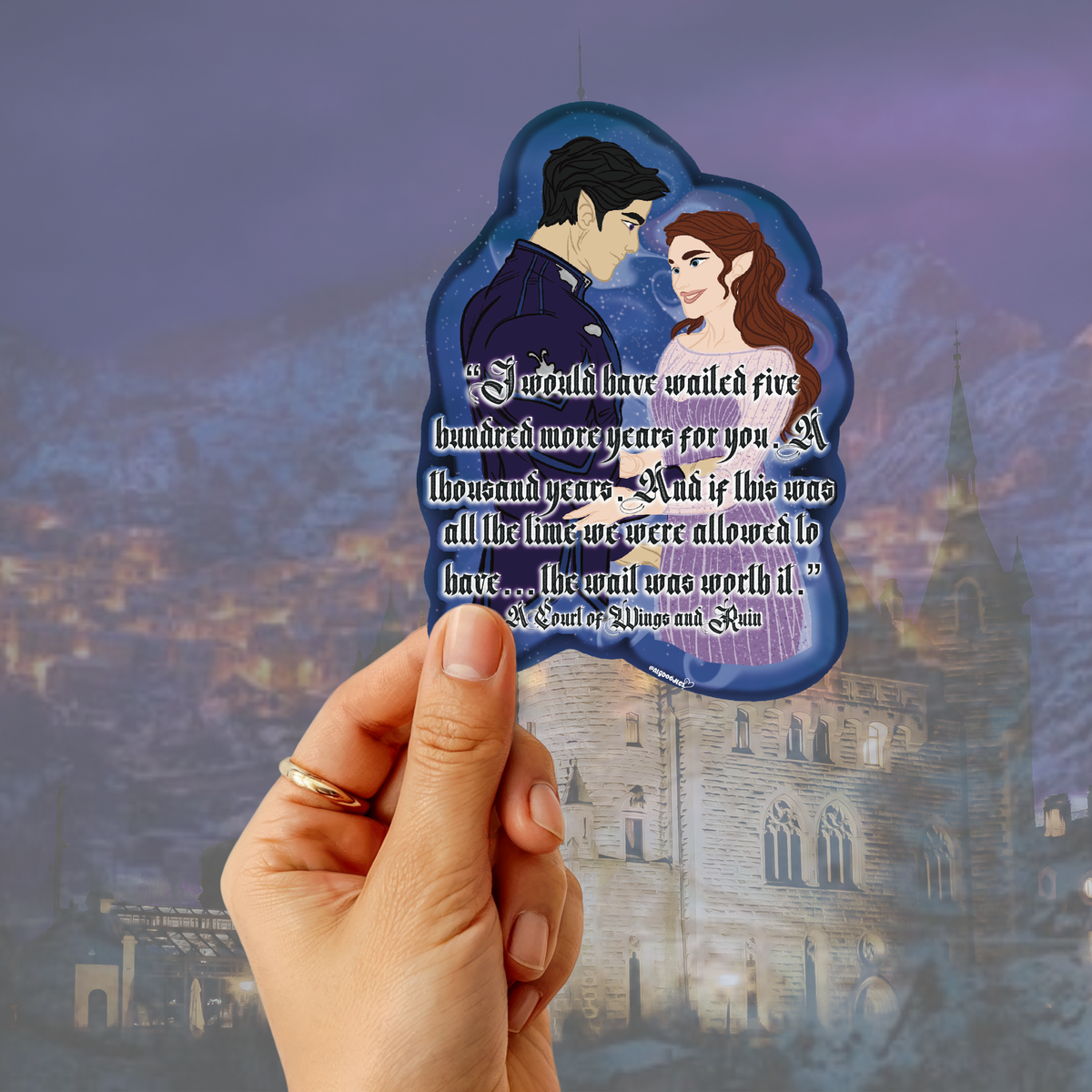 A hand holding a die-cut acrylic charm featuring an illustrated couple and a quote set against a blurred fantasy castle background.