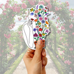 A hand holds a die-cut sticker featuring a stylized illustration of a woman with lips and a hand, with colorful flowers blooming from her body, set against a blurred garden background of pink roses.