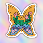 A butterfly-shaped patch or sticker featuring a colorful illustrated design with a green frog, pink flowers, blue and yellow sections, and a white border, shown against a soft pastel rainbow gradient background.