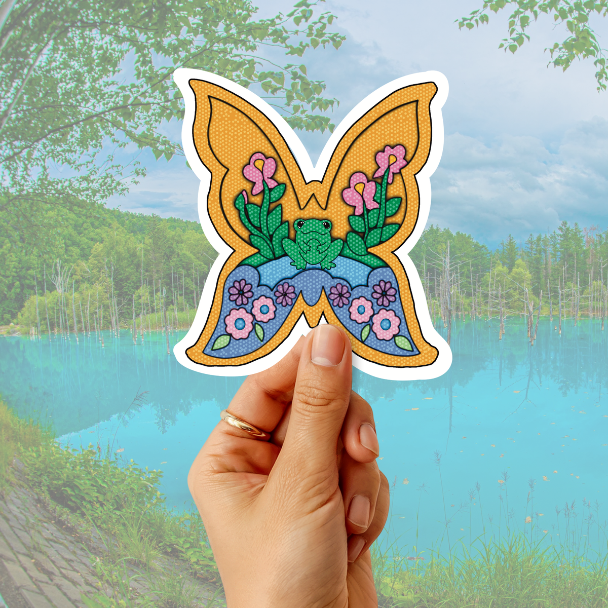A hand holds a butterfly-shaped sticker or patch with a colorful embroidered-style design featuring pink flowers, green leaves, and a blue frog motif, set against a blurred outdoor lake and forest background.