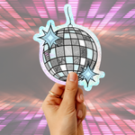 A hand holding a die-cut sticker or ornament shaped like a disco ball with two star accents, set against a pink and purple glowing grid background.