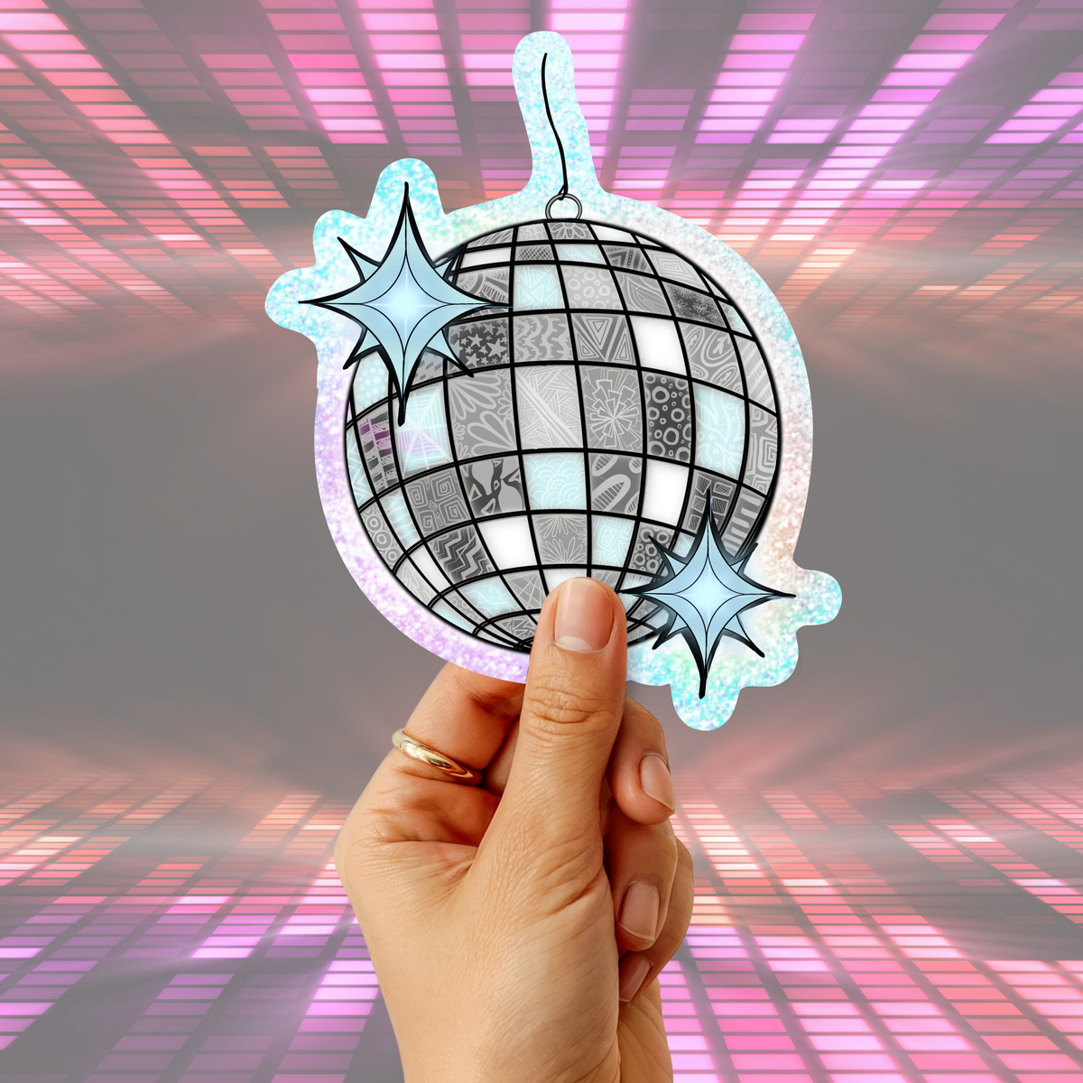 A hand holding a die-cut sticker or ornament shaped like a disco ball with two star accents, set against a pink and purple glowing grid background.
