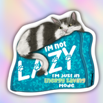 A die-cut sticker featuring a gray and white cat lounging on a teal patterned cushion with the text 'I'm not LAZY I'm just in Energy Saving Mode'. The sticker is shown against a pastel rainbow gradient background.