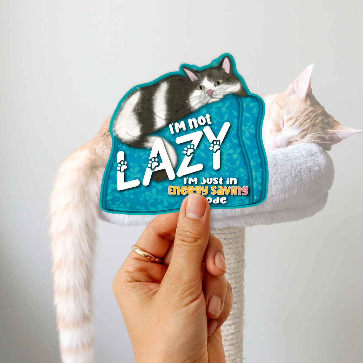 A hand holds a die-cut sticker featuring a black-and-white cat on a turquoise background with the text 'I'M NOT LAZY I'M JUST IN ENERGY SAVINGS MODE'. Behind the sticker, a real cat rests on a white cat tree against a plain light gray wall.
