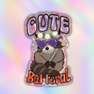 A die-cut sticker featuring a cartoon raccoon with white daisies on its head, purple heart-shaped sunglasses, and a knife, with the text 'CUTE But Feral' in stylized lettering, shown on a pastel iridescent background.