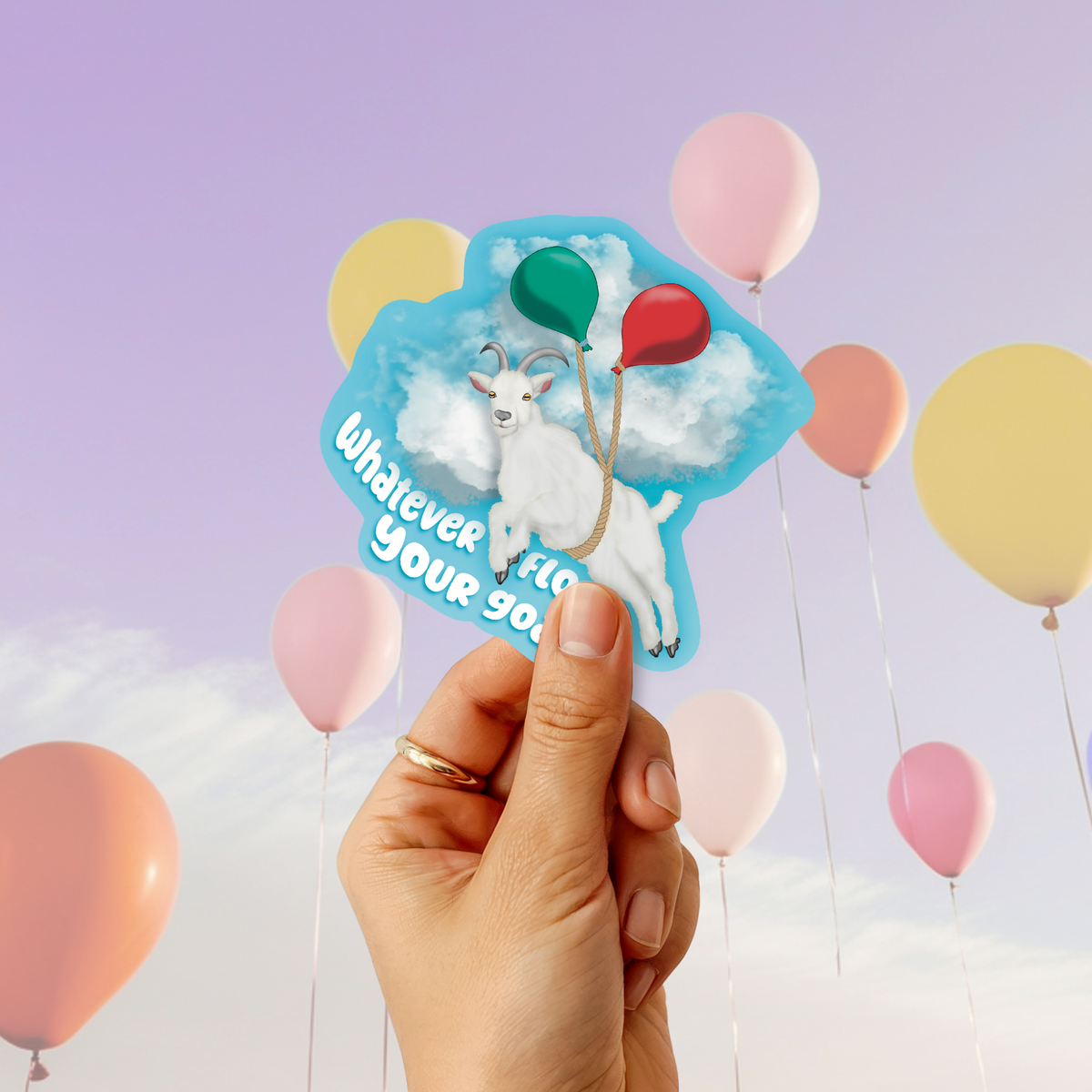A hand holds a die-cut sticker featuring a white goat on a blue cloud background with balloons and the text 'Whatever floats your boat'. The scene is set against a pastel sky with floating balloons.