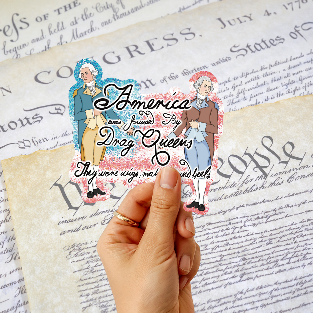 A hand holds a die-cut sticker featuring two historical figures and the text 'America was founded by Drag Queens. They wore wigs, makeup and heels .