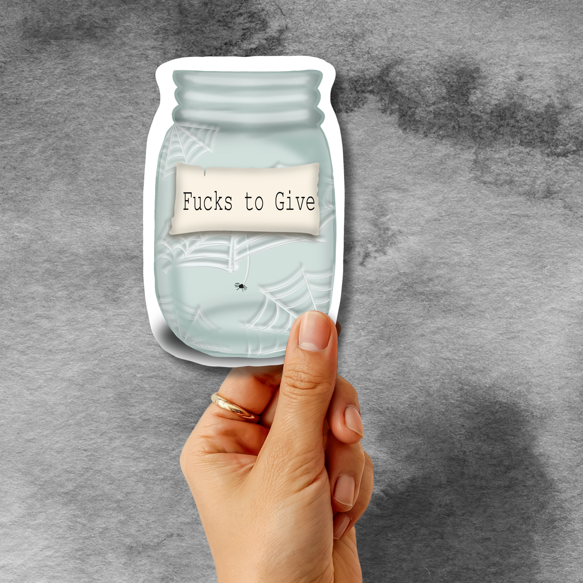A hand holds a die-cut sticker shaped like a mason jar filled with pale blue water and spiderweb graphics, with a small paper label reading 'Fucks to Give,' against a textured gray background.