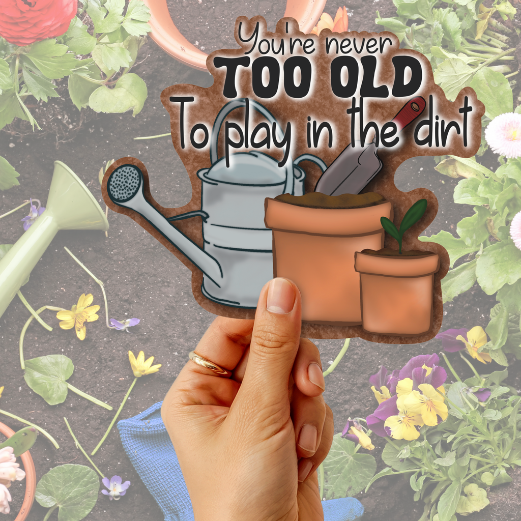 A hand holds a die-cut sticker featuring gardening tools and the quote 'You're never TOO OLD To play in the dirt' against a garden-themed background with flowers and tools.