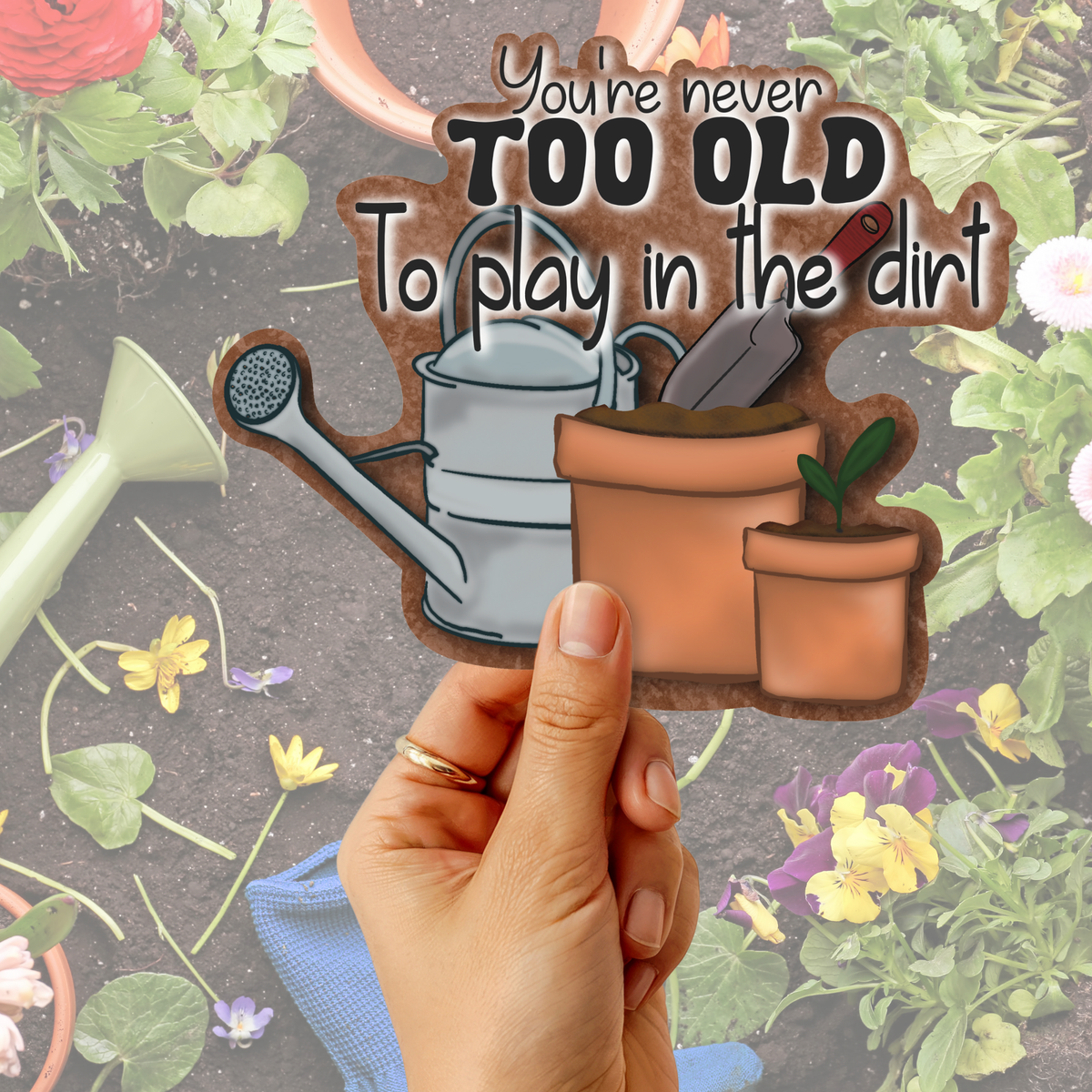 A hand holds a die-cut sticker featuring gardening tools and the quote 'You're never TOO OLD To play in the dirt' against a garden-themed background with flowers and tools.