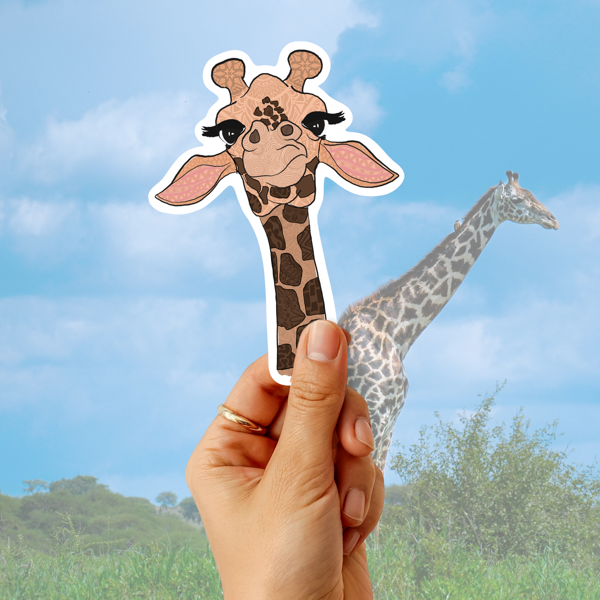 A hand holds a die-cut sticker of a cute cartoon giraffe head in front of a real giraffe photo against a blue sky and grass background.