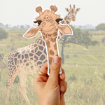A hand holds a die-cut sticker of a cute, long-eared giraffe in front of a blurred background photo of a real giraffe in a savanna.