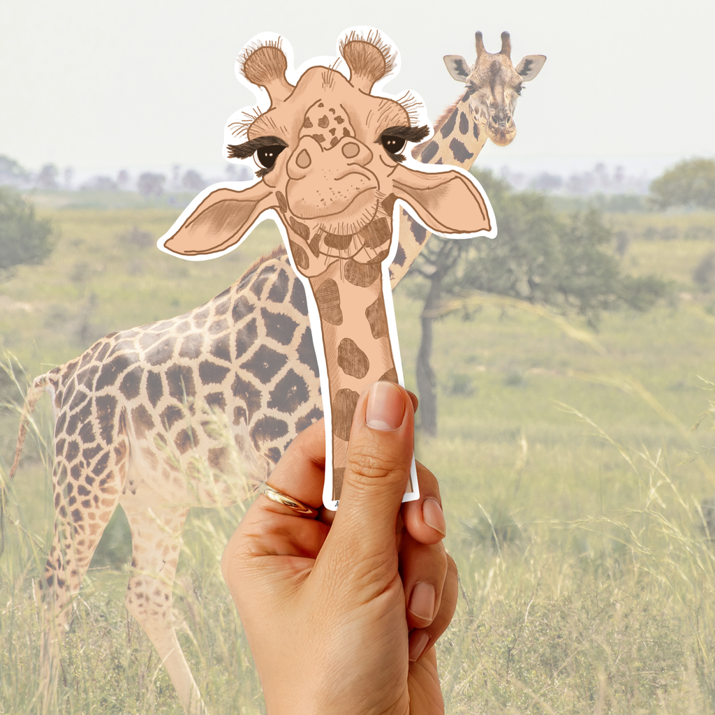 A hand holds a die-cut sticker of a cute, long-eared giraffe in front of a blurred background photo of a real giraffe in a savanna.