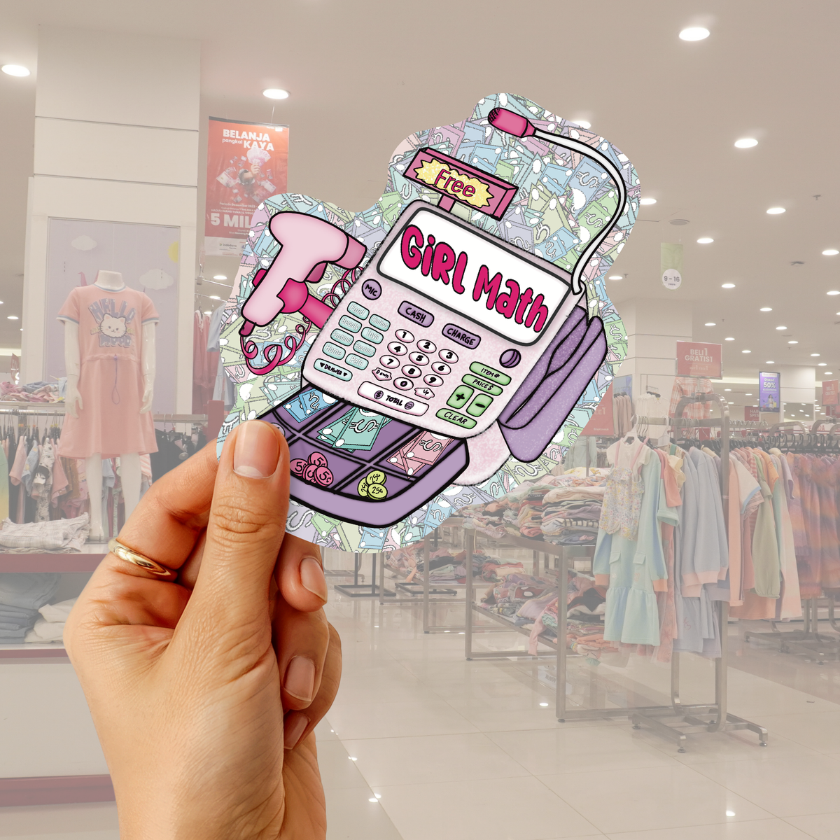 A hand holds a die-cut sticker featuring a retro calculator with the text 'GIRL MATH' and a pink phone, set against a blurred clothing store background.