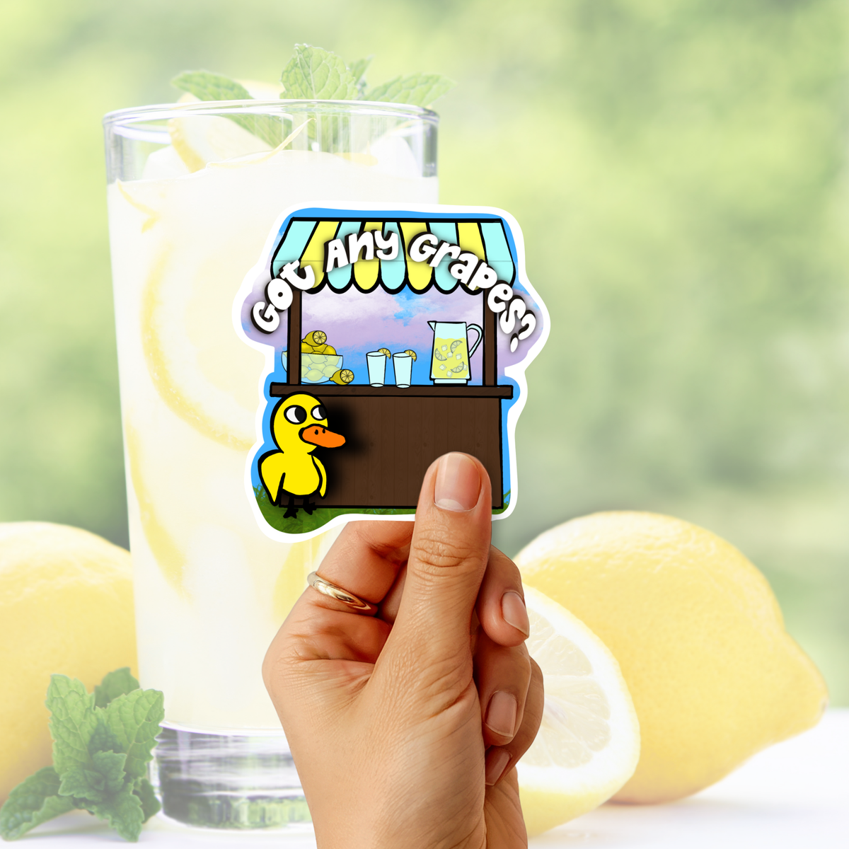 A hand holds a die-cut sticker featuring a cartoon yellow duck and a lemonade stand with the text 'God Any Grapes' against a blurred background of a glass of lemonade and lemons.