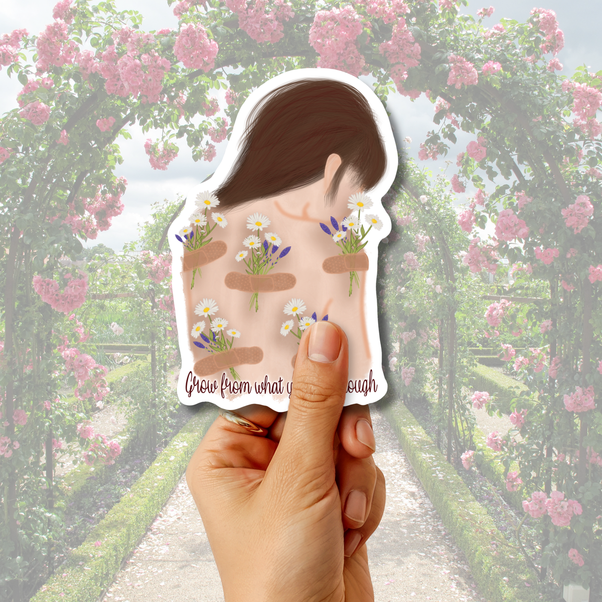 A hand holds a die-cut sticker featuring an illustration of a woman's back with flowers in her hair and the text 'Grew from what was enough', set against a blurred garden archway background of pink flowers.