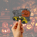 A hand holds a die-cut sticker featuring a yellow car with a skull emblem and a large orange jack-o'-lantern, with the text 'GET IN LOOSER WE ARE SAYING HALLOWEENTOWN' over a blurred Halloween yard scene filled with glowing pumpkins.