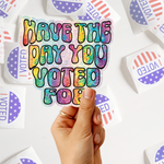 A hand holds a die-cut sticker with the text 'HAVE THE DAY YOU VOTED FOR' in a colorful rainbow glitter pattern. The background is filled with other stickers, including circular 'I VOTED' stickers with red and white stripes and blue stars.