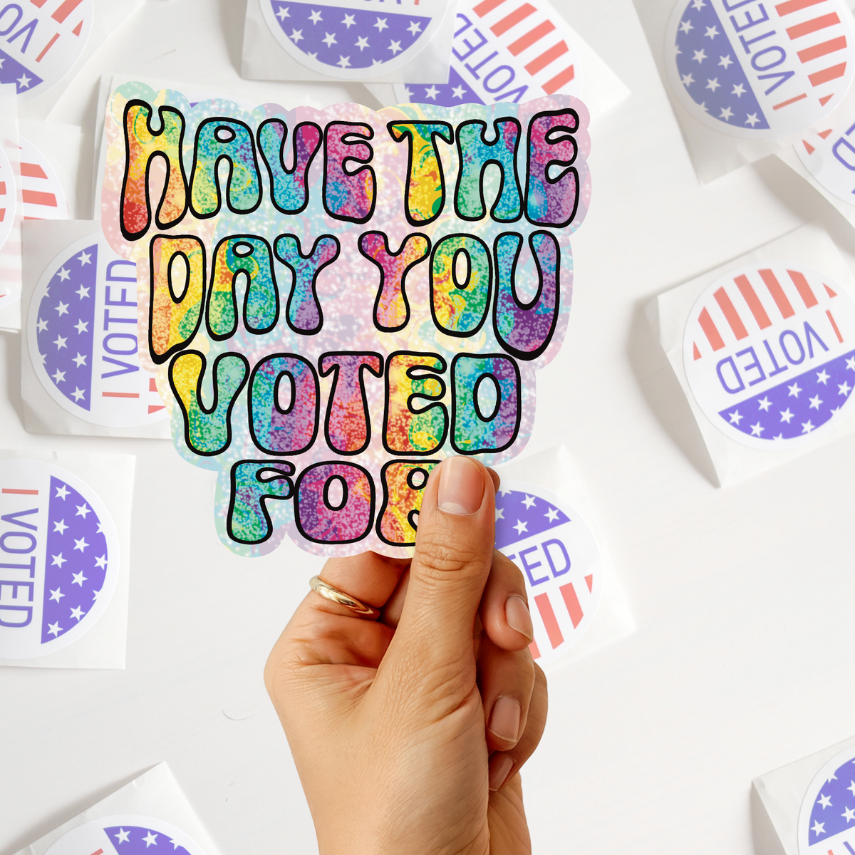 A hand holds a die-cut sticker with the text 'HAVE THE DAY YOU VOTED FOR' in a colorful rainbow glitter pattern. The background is filled with other stickers, including circular 'I VOTED' stickers with red and white stripes and blue stars.