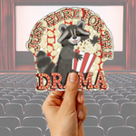 A hand holds a die-cut sticker featuring a raccoon holding a popcorn bucket with the text 'JUST HERE FOR THE DRAMA' against a blurred movie theater audience background.