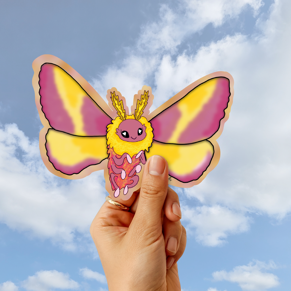 A hand holds a die-cut sticker of a cute cartoon moth against a blue sky with clouds background.