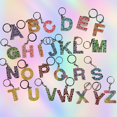 Alphabet letter keychains arranged in rows on a pastel rainbow gradient background. Each letter charm is decorated with small colorful polka dots and has a split key ring attached.