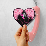 A hand holds a heart-shaped sticker featuring a black hooded ghost-face character on a pink background with hearts and the words 'SUITABLE' and 'LOVE'. A pink corded telephone is partially visible behind it against a light gray textured wall.