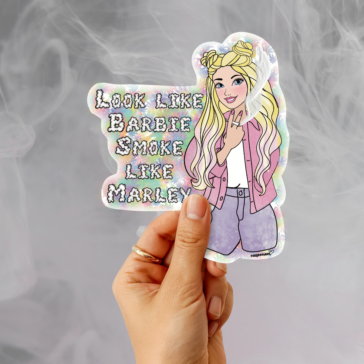 A hand holds a die-cut sticker featuring an illustrated blonde woman in a pink jacket and lavender pants next to the text 'LOOK LIKE BARBIE SMOKE LIKE MARLEY' in pastel rainbow letters, against a smoky gray background.