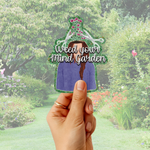 A hand holds a die-cut sticker featuring an illustrated figure with a purple shirt, brown braid, pink flowers, and the text 'Weed your Mind Garden' against a blurred garden background.
