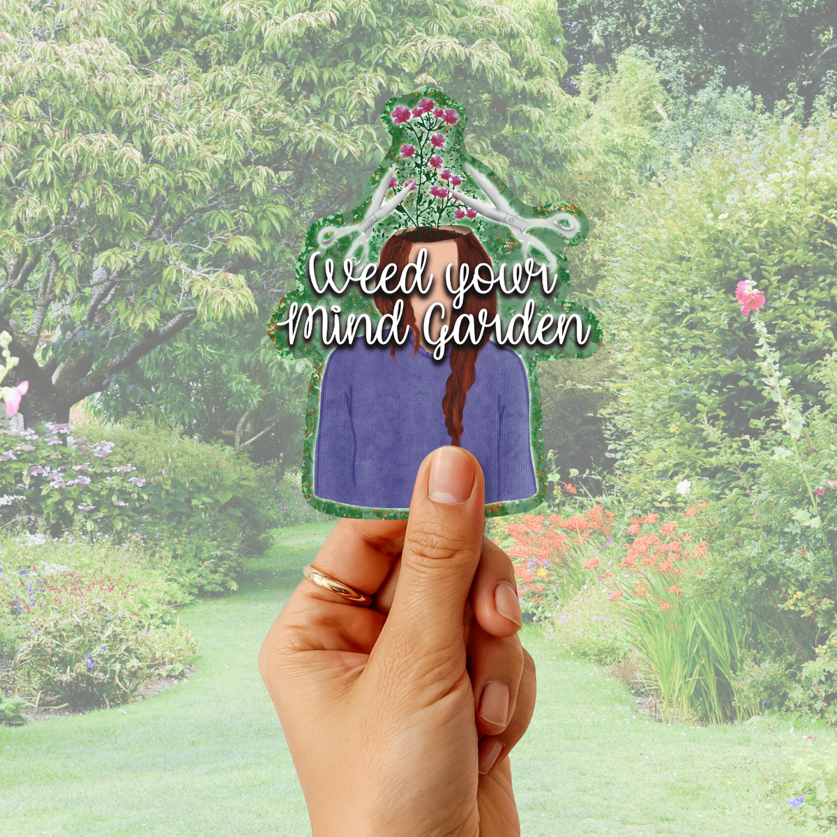 A hand holds a die-cut sticker featuring an illustrated figure with a purple shirt, brown braid, pink flowers, and the text 'Weed your Mind Garden' against a blurred garden background.