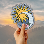 A hand holds a die-cut sticker featuring a yellow sun and white crescent moon with a blue-gray background and a pink flower, set against a misty mountain landscape at sunset.