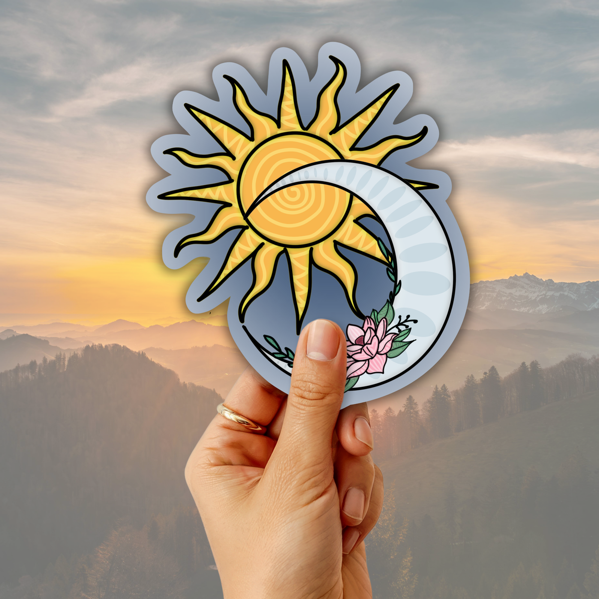 A hand holds a die-cut sticker featuring a yellow sun and white crescent moon with a blue-gray background and a pink flower, set against a misty mountain landscape at sunset.