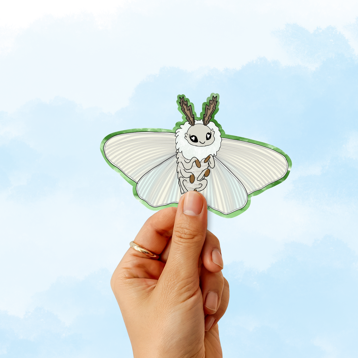 A hand holding a die-cut sticker of a cute cartoon moth with white wings and a green outline, set against a light blue watercolor sky background.