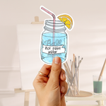A hand holds a die-cut sticker shaped like a mason jar filled with blue liquid, featuring a pink striped straw, a yellow lemon slice, and the text 'Ball Not paint water' on the label. The background shows a softly blurred art studio setting with an easel, paintbrushes, and a small glass of green paint.