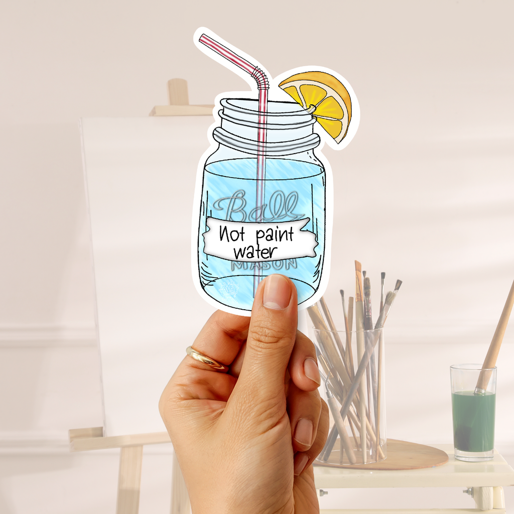 A hand holds a die-cut sticker shaped like a mason jar filled with blue liquid, featuring a pink striped straw, a yellow lemon slice, and the text 'Ball Not paint water' on the label. The background shows a softly blurred art studio setting with an easel, paintbrushes, and a small glass of green paint.