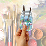 A hand holds a die-cut sticker shaped like a paint jar with two paintbrushes and the text 'Paint water Do not drink'. The background is a soft, painterly multicolor scene with paintbrushes and paint pots.