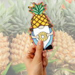 A hand holds a die-cut sticker shaped like a cartoon gnome wearing a pineapple body and holding a pineapple slice, with a blurred background of many pineapples.