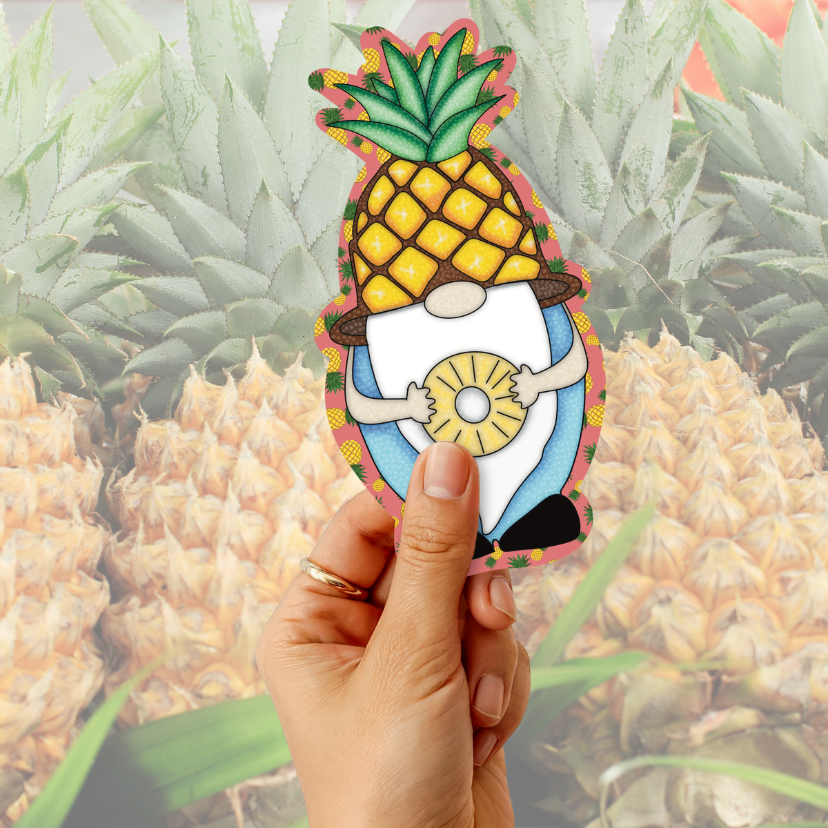 A hand holds a die-cut sticker shaped like a cartoon gnome wearing a pineapple body and holding a pineapple slice, with a blurred background of many pineapples.