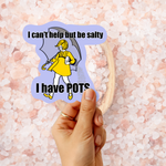 A hand holds a die-cut sticker featuring a cartoon woman in a yellow raincoat and umbrella, with the text 'I can't help but be salty' at the top and 'I have POTS' at the bottom. The background is filled with pink salt crystals.