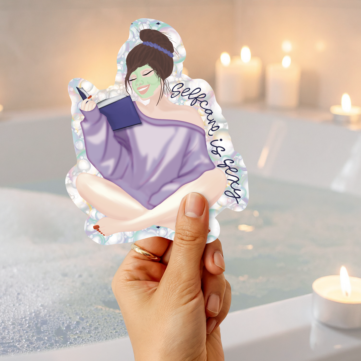 A hand holds a die-cut sticker featuring an illustrated woman in a purple robe and green face mask with the text 'Believe is enough', set against a blurred bathroom scene with candles and a bathtub.