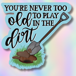 Decorative sign with a shovel and text 'You're never too old to play in the dirt' on a colorful background.