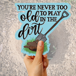 A hand holds a die-cut sticker that reads 'YOU'RE NEVER TOO OLD TO PLAY IN THE DIRT' with a shovel illustration, against a sandy outdoor ground background.