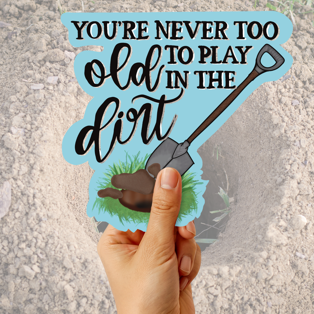 A hand holds a die-cut sticker that reads 'YOU'RE NEVER TOO OLD TO PLAY IN THE DIRT' with a shovel illustration, against a sandy outdoor ground background.