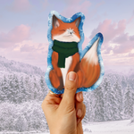 A hand holding a die-cut sticker or card featuring an illustrated orange fox wearing a green scarf, set against a snowy forest landscape background.