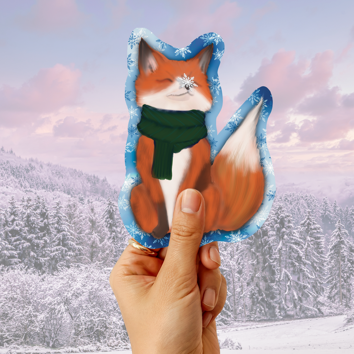 A hand holding a die-cut sticker or card featuring an illustrated orange fox wearing a green scarf, set against a snowy forest landscape background.