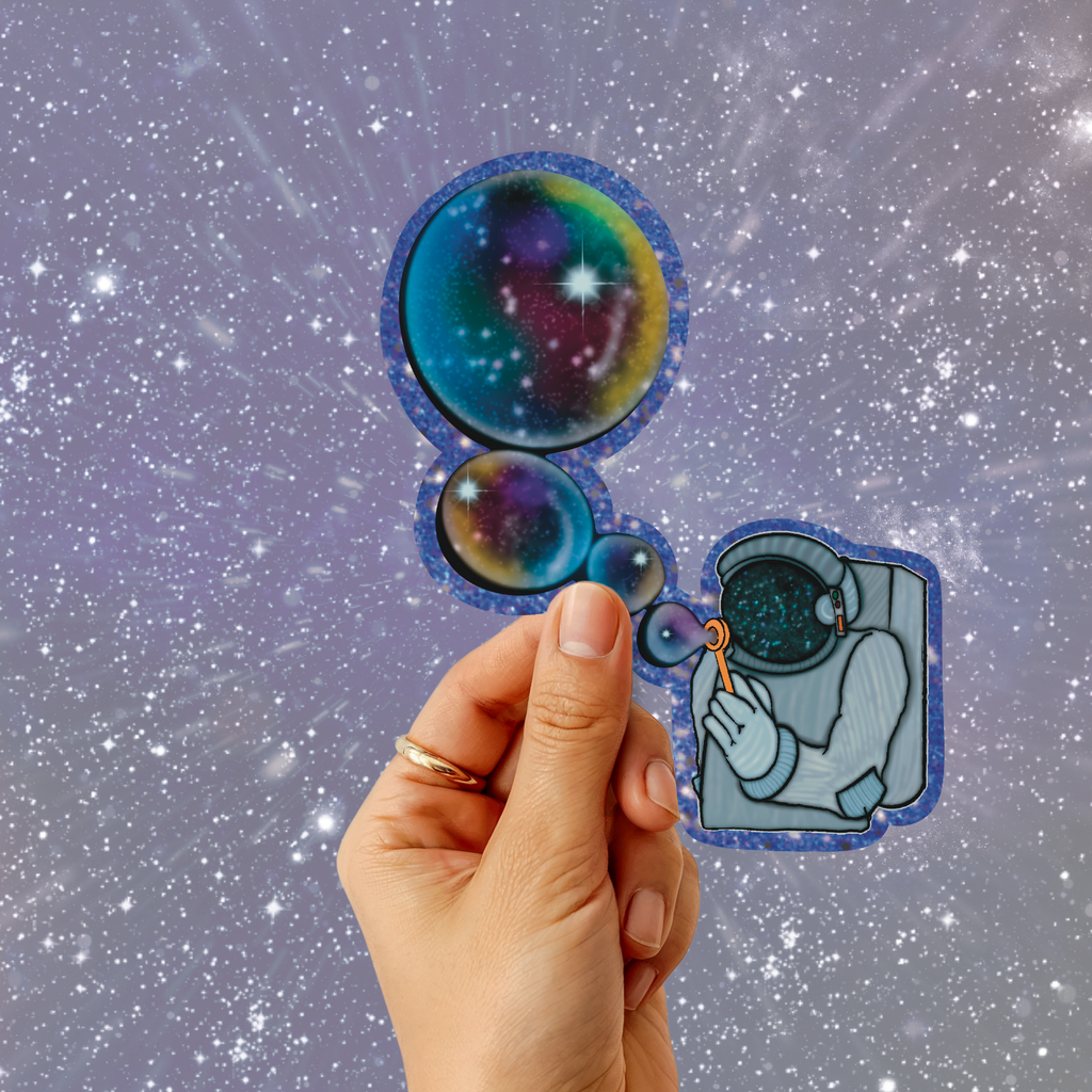 A hand holds two die-cut stickers against a starry space background. One sticker shows a colorful galaxy bubble, and the other shows an astronaut with a galaxy helmet.