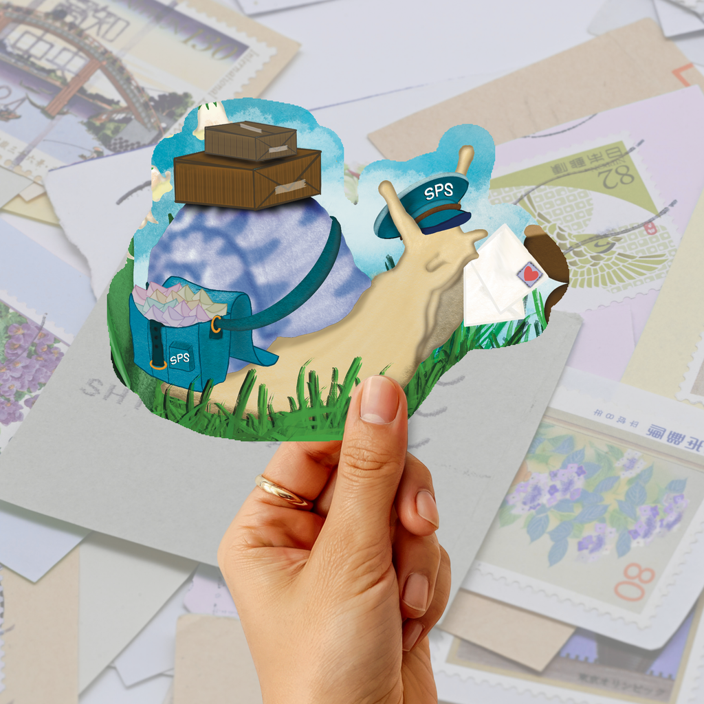 A hand holds a die-cut sticker or pop-up card featuring a whimsical postage stamp snail illustration with a blue backpack, brown boxes, green grass, and a light blue cloud background. Scattered postage stamps fill the soft-focus background.