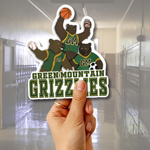 A hand holds a die-cut sticker featuring three cartoon grizzlies in green and gold uniforms with the text 'GREEN MOUNTAIN GRIZZLES' and 'GM'. The background shows a blurred school hallway.