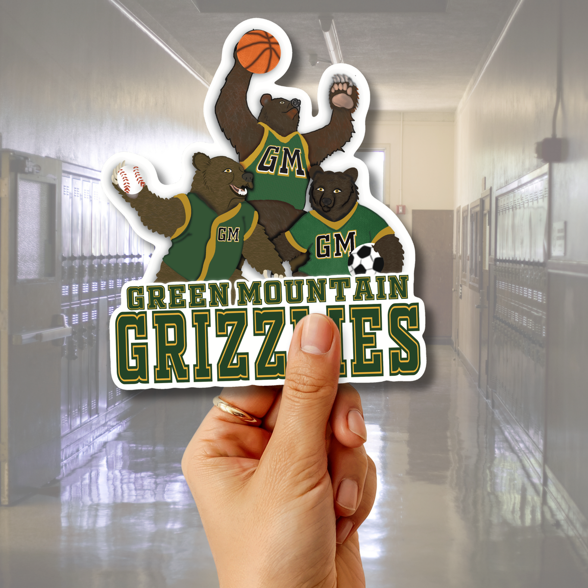 A hand holds a die-cut sticker featuring three cartoon grizzlies in green and gold uniforms with the text 'GREEN MOUNTAIN GRIZZLES' and 'GM'. The background shows a blurred school hallway.
