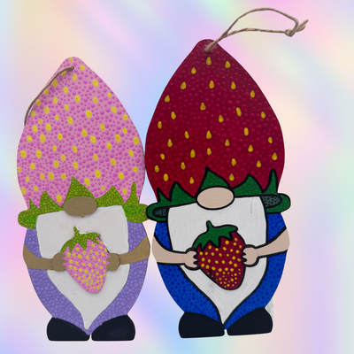 Two decorative gnome ornaments shaped like strawberries, shown side by side against a soft pastel rainbow background. One gnome has a pink and purple top with yellow dots, green accents, and holds a pink strawberry.